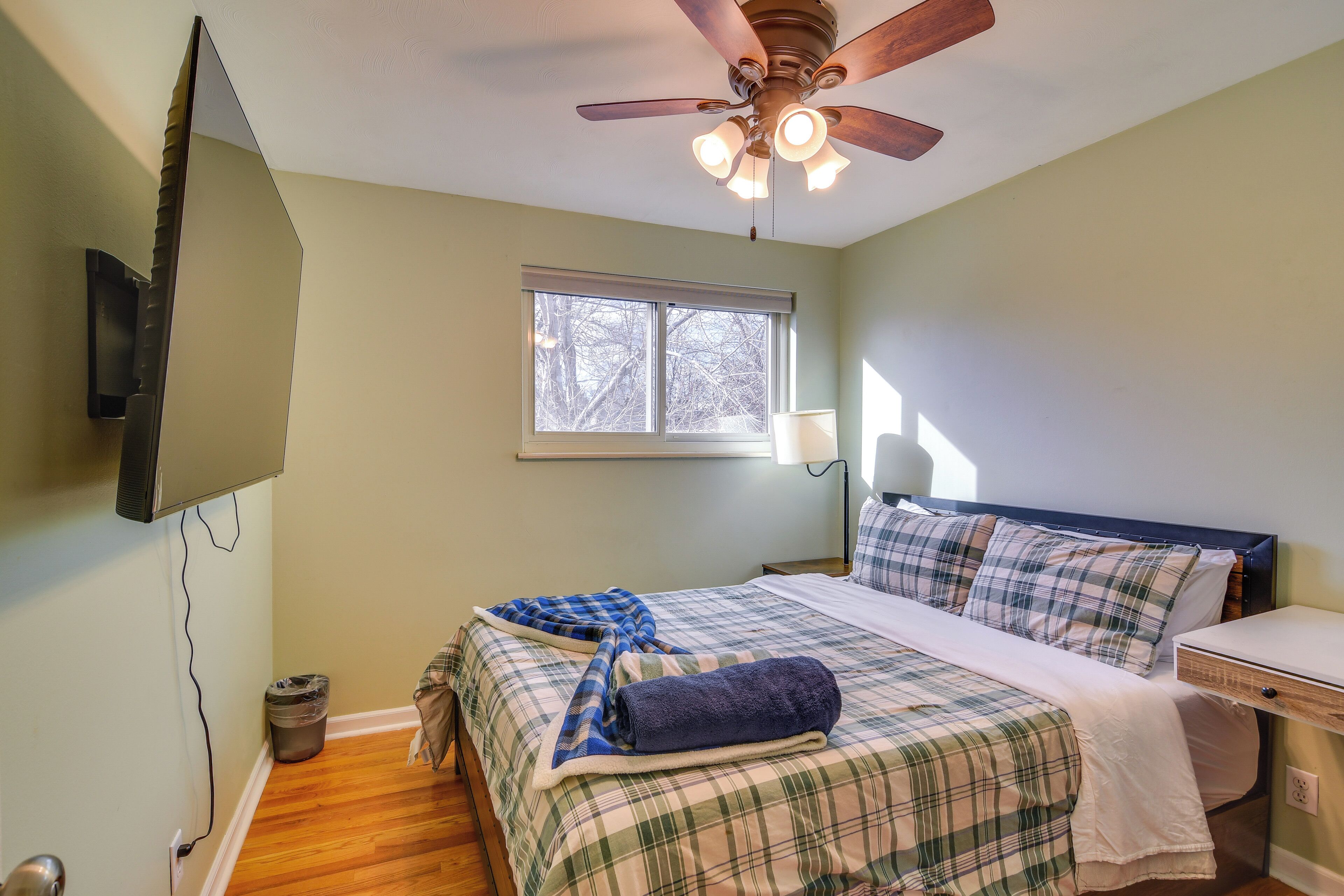 A bedroom with a bed, a ceiling fan, a window, and a TV mounted on the wall.