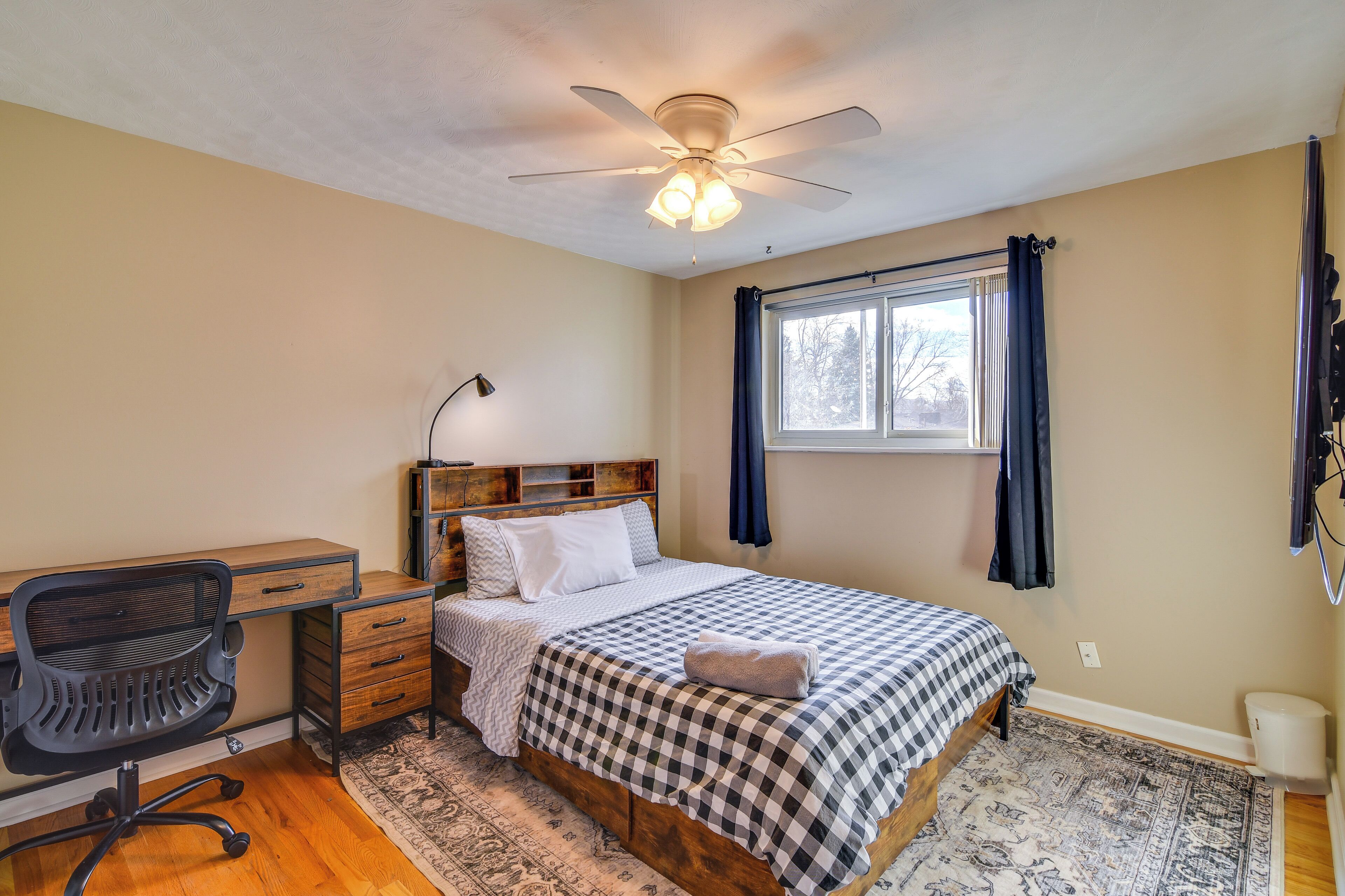 Une chambre à coucher comprenant un lit, un bureau, une chaise, un ventilateur de plafond, une fenêtre et des rideaux.