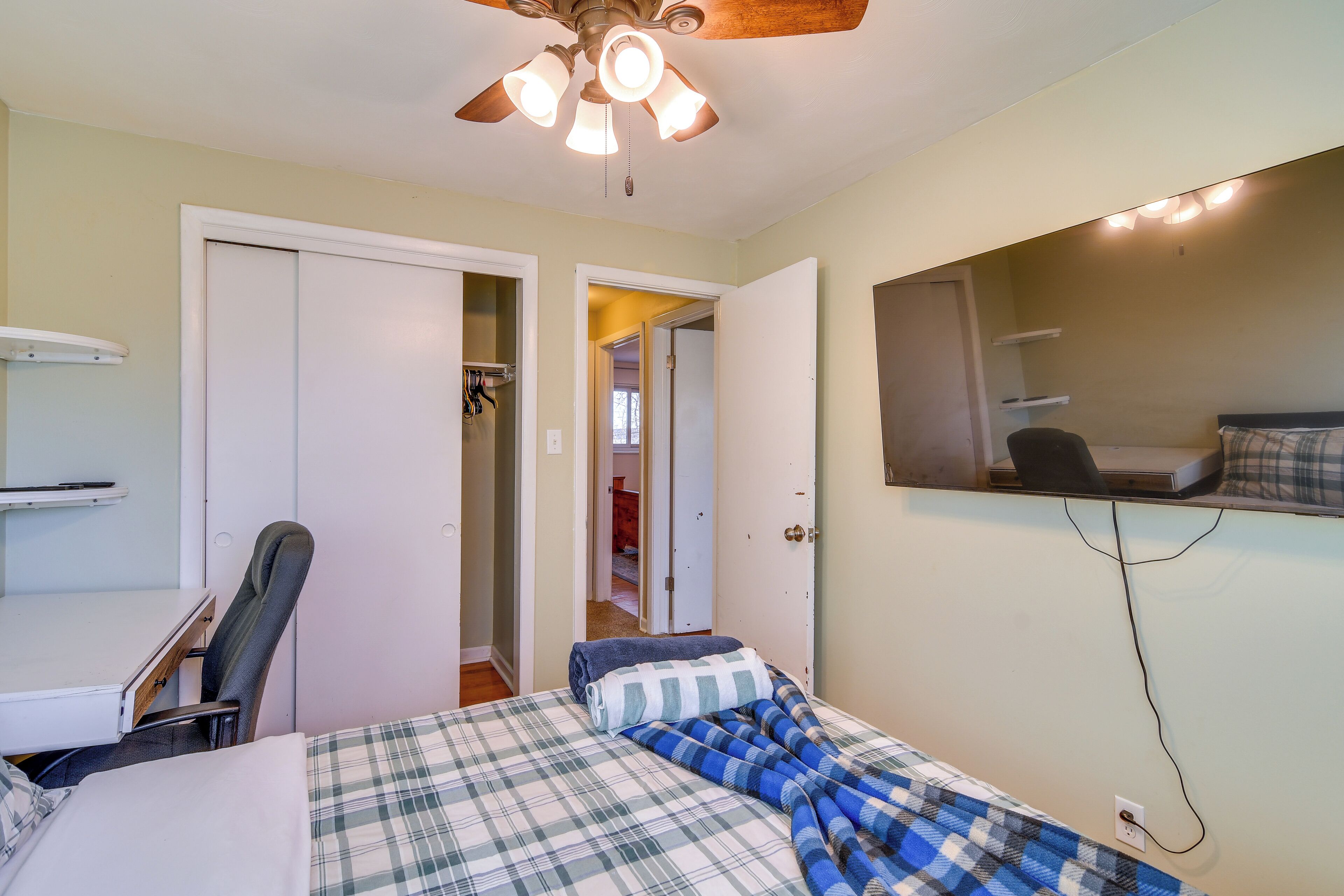 A bedroom with a bed, a desk, a chair, a ceiling fan, and a mirror.