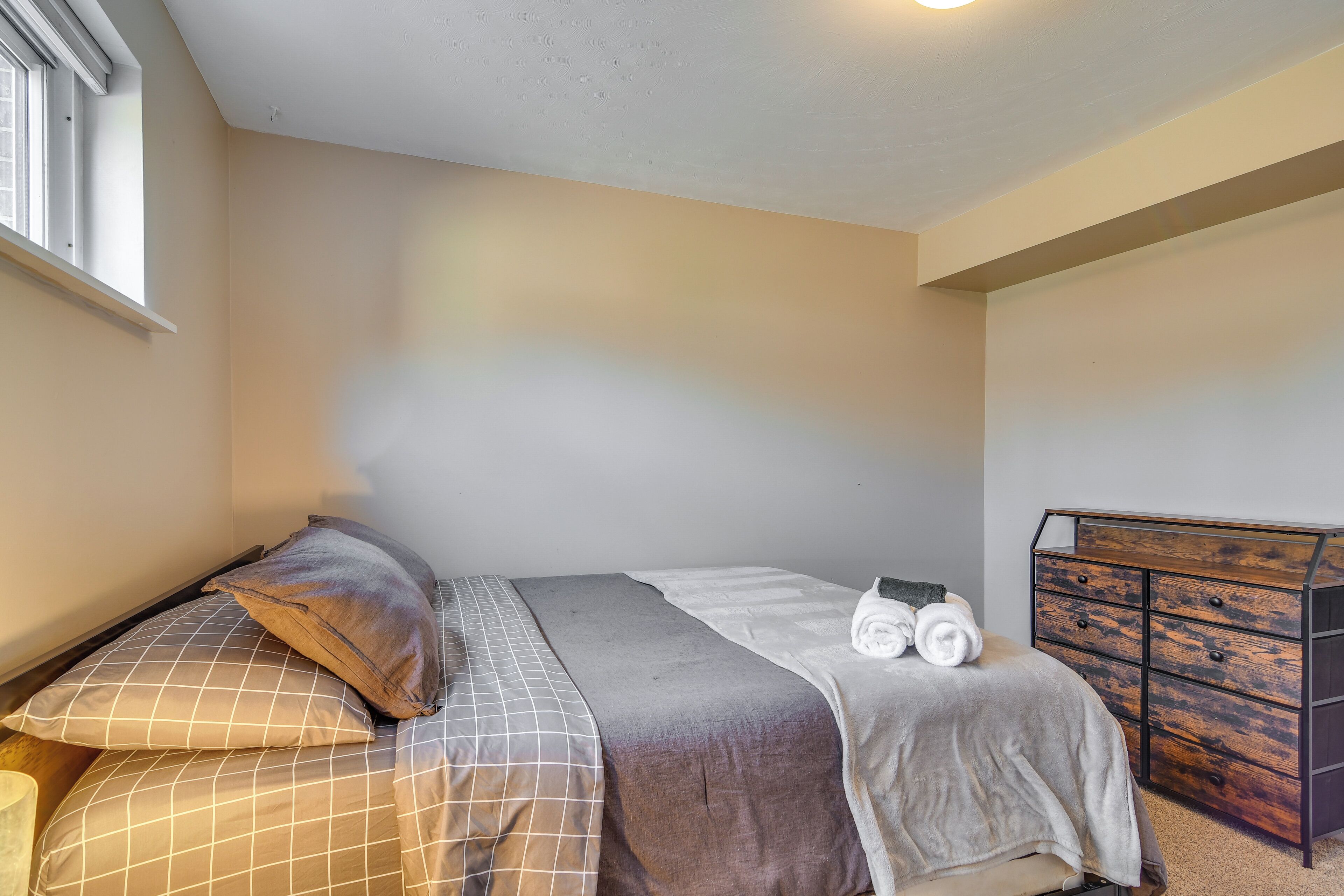 Een slaapkamer met een bed, kussens, een deken, een houten dressoir en een raam.