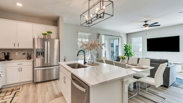 A modern kitchen with stainless steel appliances, a white island, and a dining area with a gray sofa and white chairs. - Reunion Resort: Brand-new Home Near Disney! (Kissimmee)