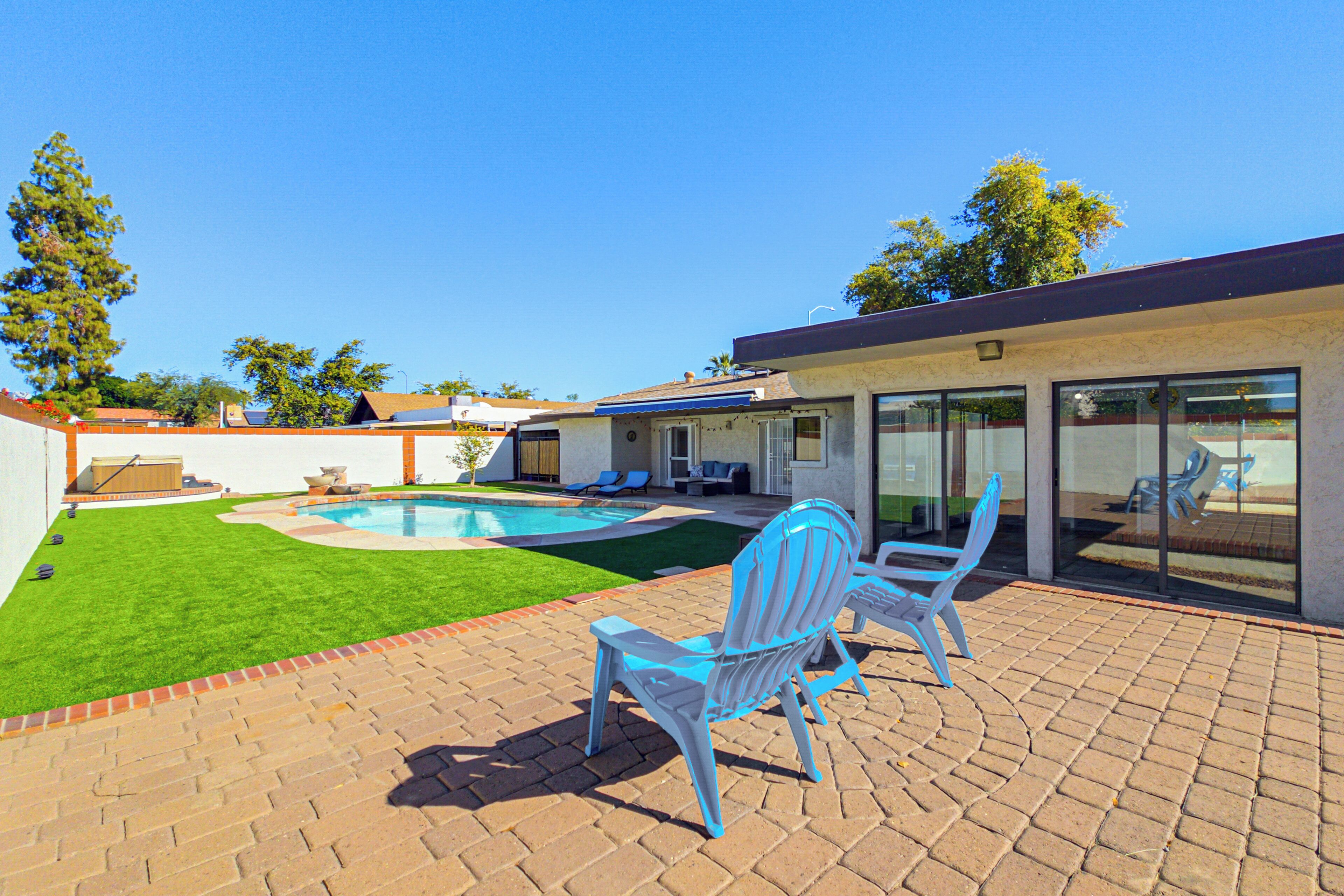 A backyard with a swimming pool, two blue Adirondack chairs, and a patio area with brick flooring.