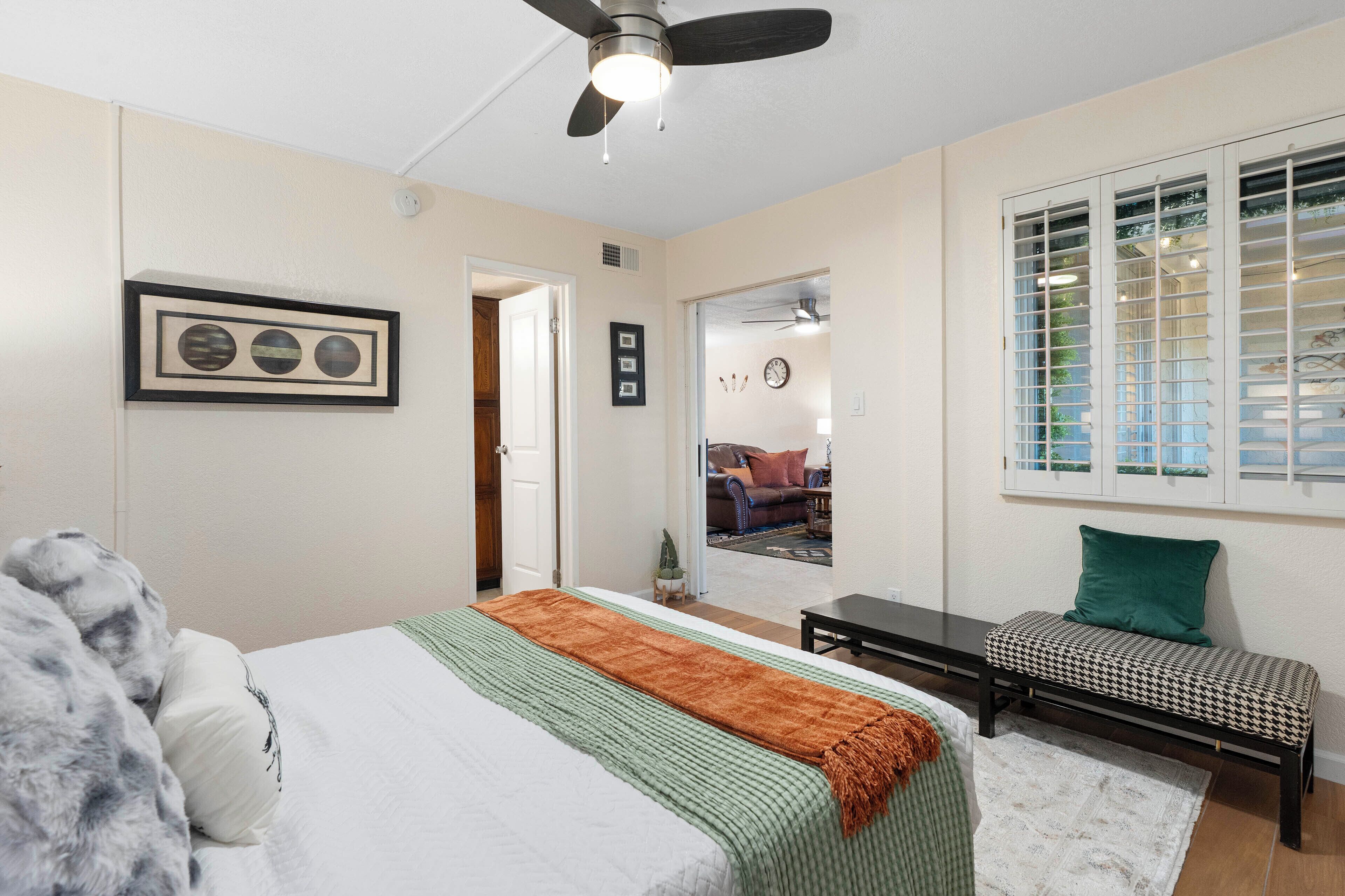 A bedroom with a bed, a ceiling fan, a window with blinds, a small table, and a bench.