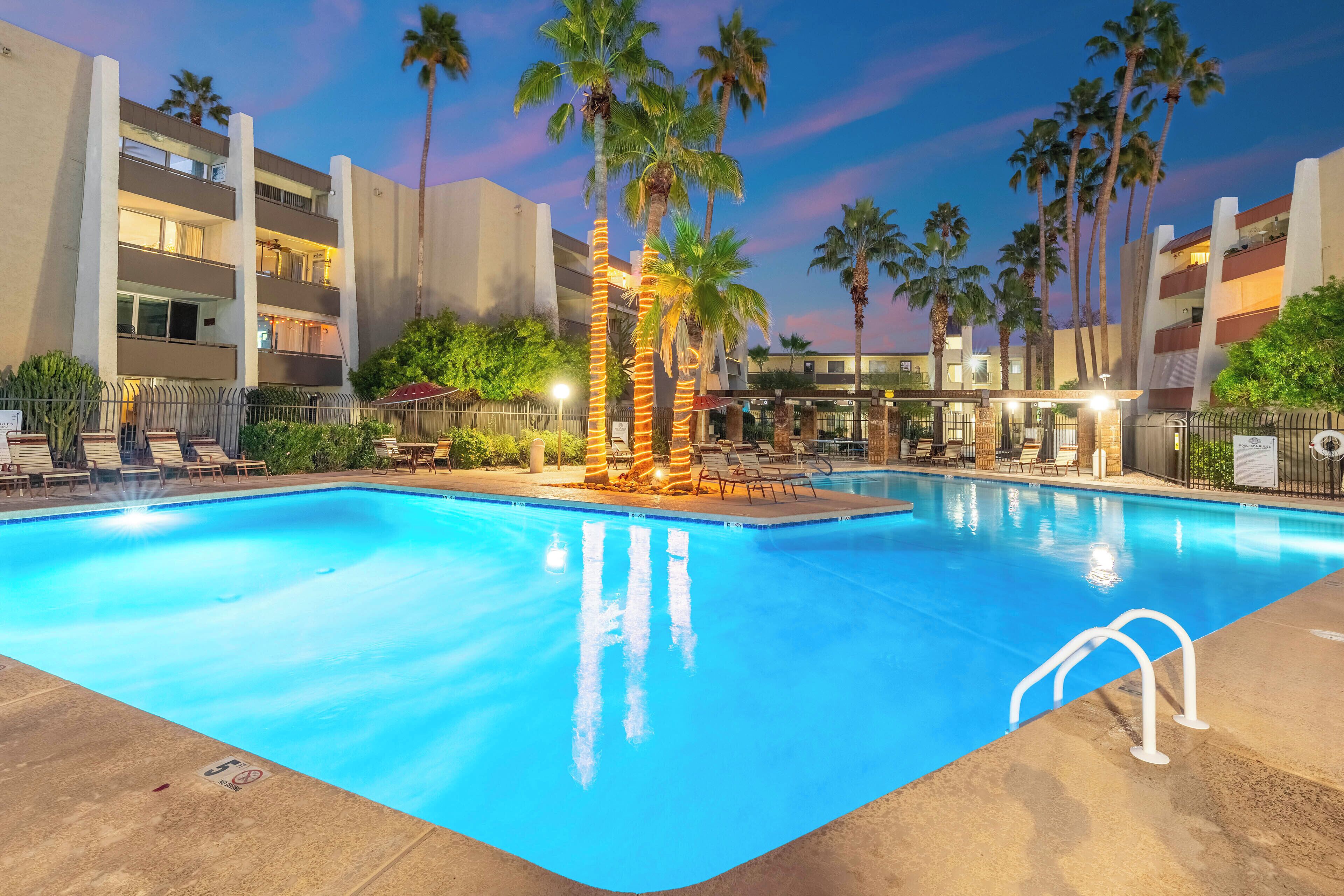 A swimming pool with palm trees and lounge chairs, surrounded by a multi-story building with balconies.