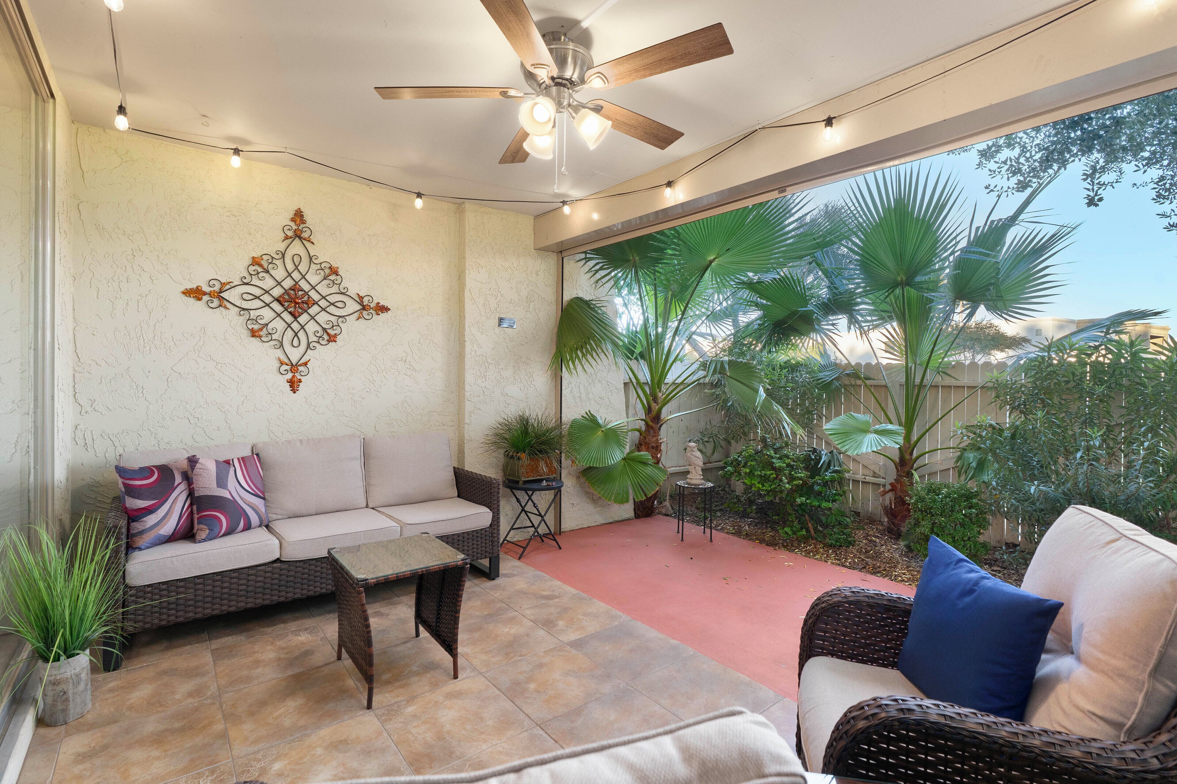 Un patio couvert doté d’un ventilateur de plafond, d’un canapé avec des coussins à motifs, d’une table basse et de plantes en pot.