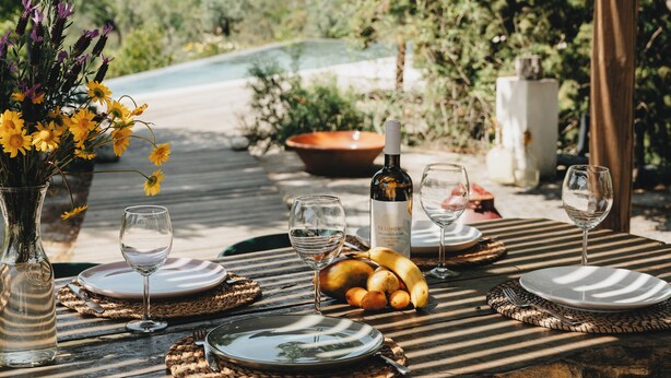 Eine Holzterrasse mit Esstisch, umgeben von Grünpflanzen und einem Pool im Hintergrund.