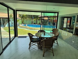 Zona de menjador amb taula de vidre i cadires de vimetla, amb vista a una piscina i un jardí.
