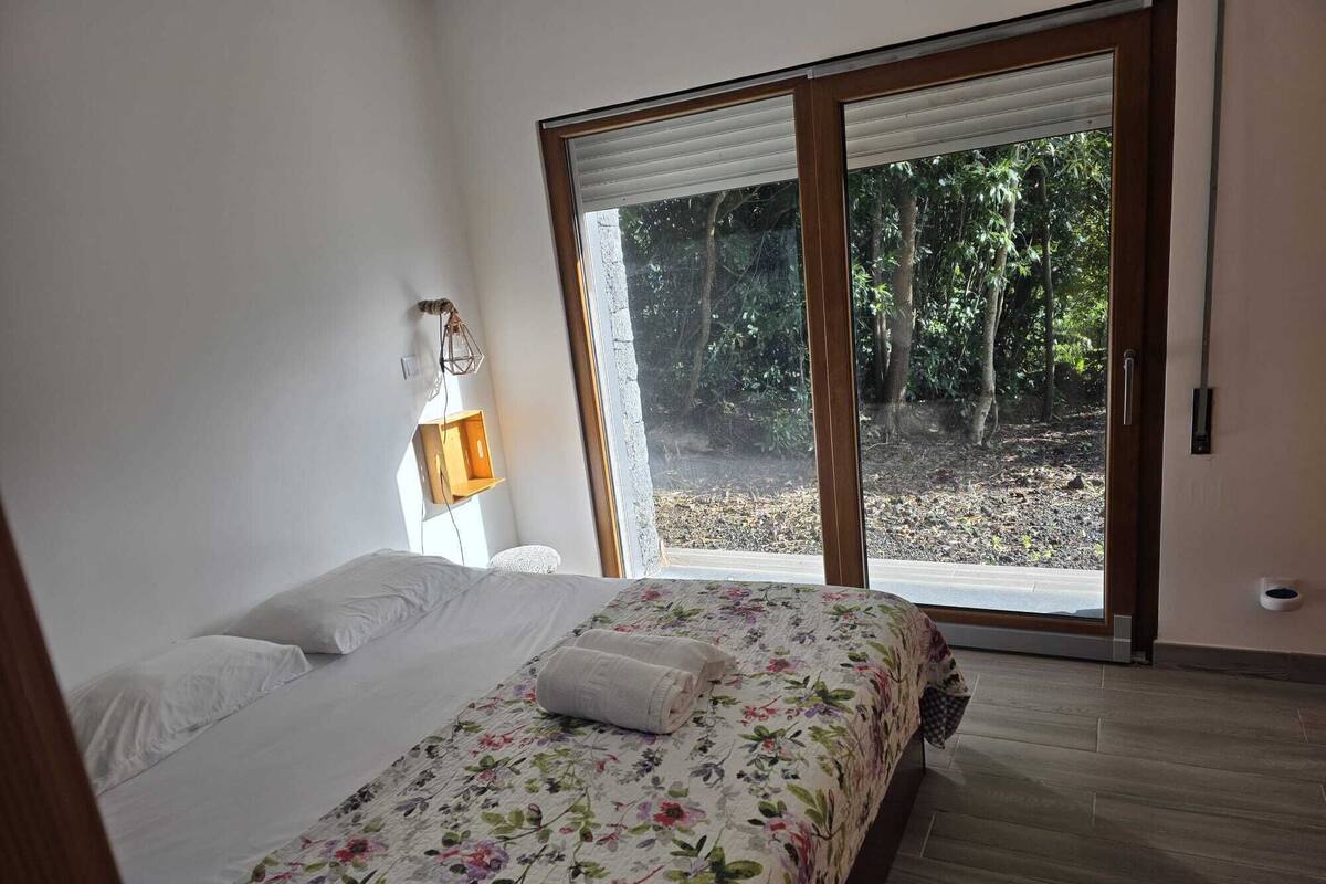 Un dormitorio con una cama, una mesita de noche, una lámpara y un ventanal corredero que da a un jardín.