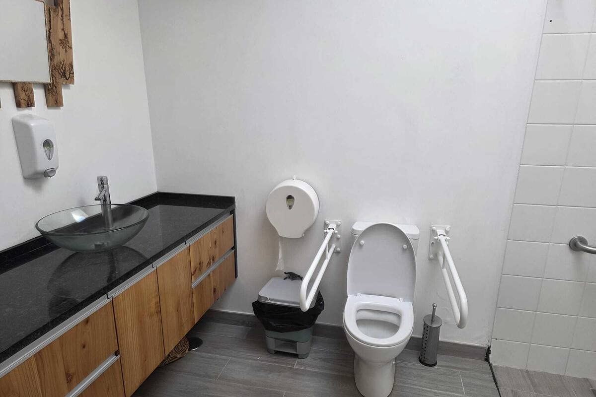 Un baño moderno con un lavabo de vidrio, tocador de madera, dos inodoros y un secador de manos.