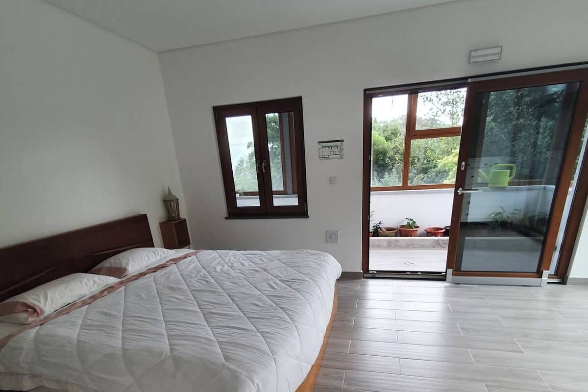 Un dormitorio con cama, ventana, puerta que da a un balcón y una mesita de noche con lámpara.