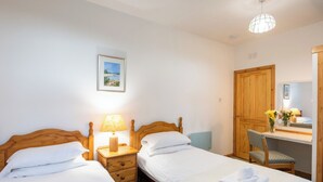 Une chambre avec deux lits simples, une table de chevet avec une lampe, une armoire en bois, un bureau avec une chaise et un miroir.