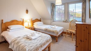 Une chambre avec deux lits simples, un bureau en bois, une chaise, une lampe et une fenêtre donnant sur des arbres.