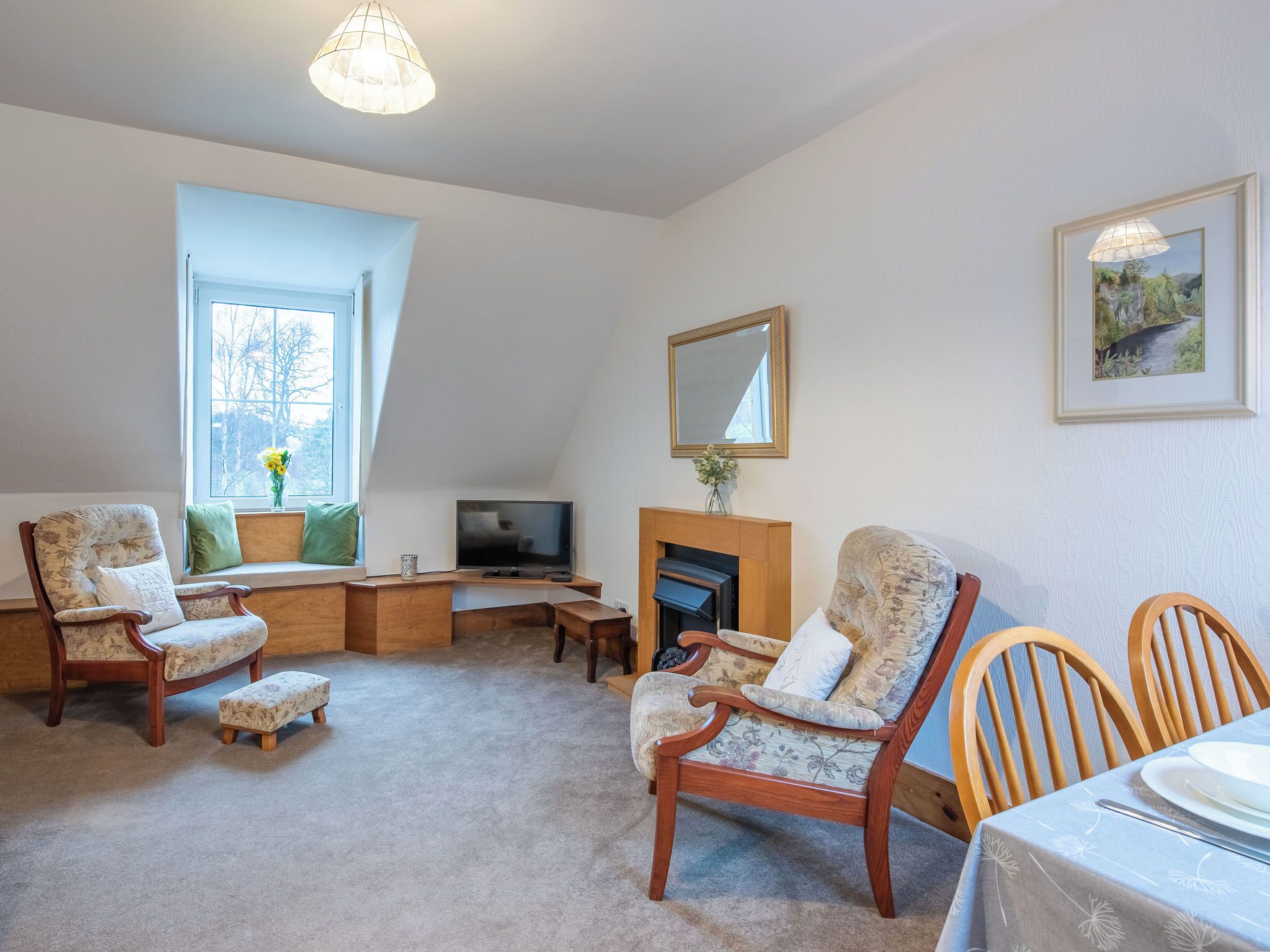 A cozy living room with a fireplace, comfortable seating, a television, and a large window with a view of trees.