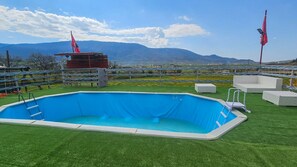 Una piscina rectangular sobre césped artificial con una escalera, rodeada por una valla y banderas, con montañas al fondo.