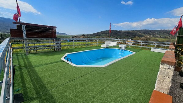 Una piscina al aire libre rodeada por una zona vallada con césped artificial, una estructura de madera y vistas a colinas al fondo.