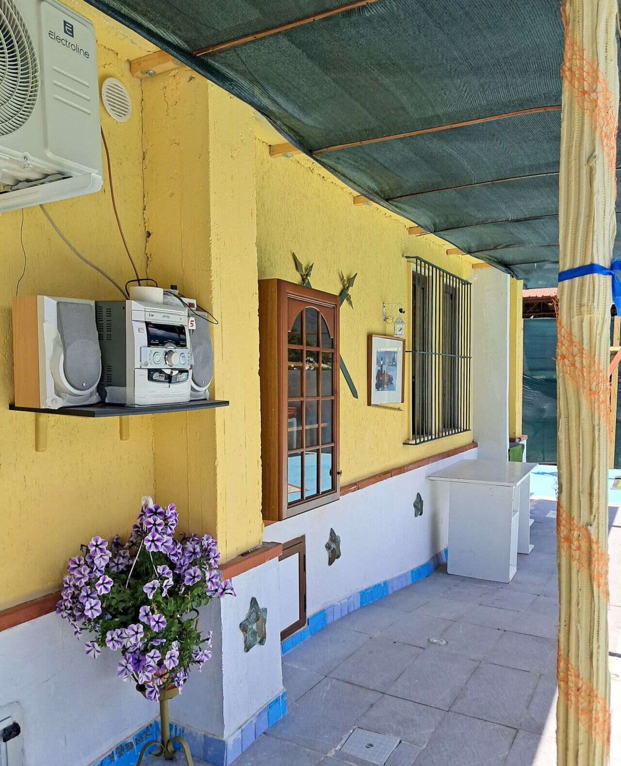A yellow building with a covered outdoor area, a wooden window, and a wall-mounted air conditioning unit.