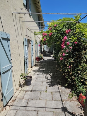 A narrow alleyway with a paved path, a building with blue shutters, and a flowering bush. - Stunning 1819 Mansion with a Pool for 14 People (Vailhourles)