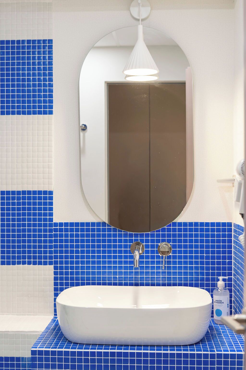 Een badkamer met een wit wastafelmeubel, blauwe tegels, een spiegel en een witte hanglamp.