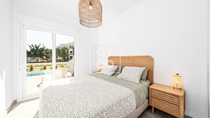 Een slaapkamer met een bed, nachtkastje, hanglamp en uitzicht op een zwembad en groen buiten.