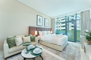 A hotel room with a bed, a sofa, a dining table with plates and glasses, and a large window with a view of buildings. - PVH - Seven Palm - Studio Beach Access (Dubai)