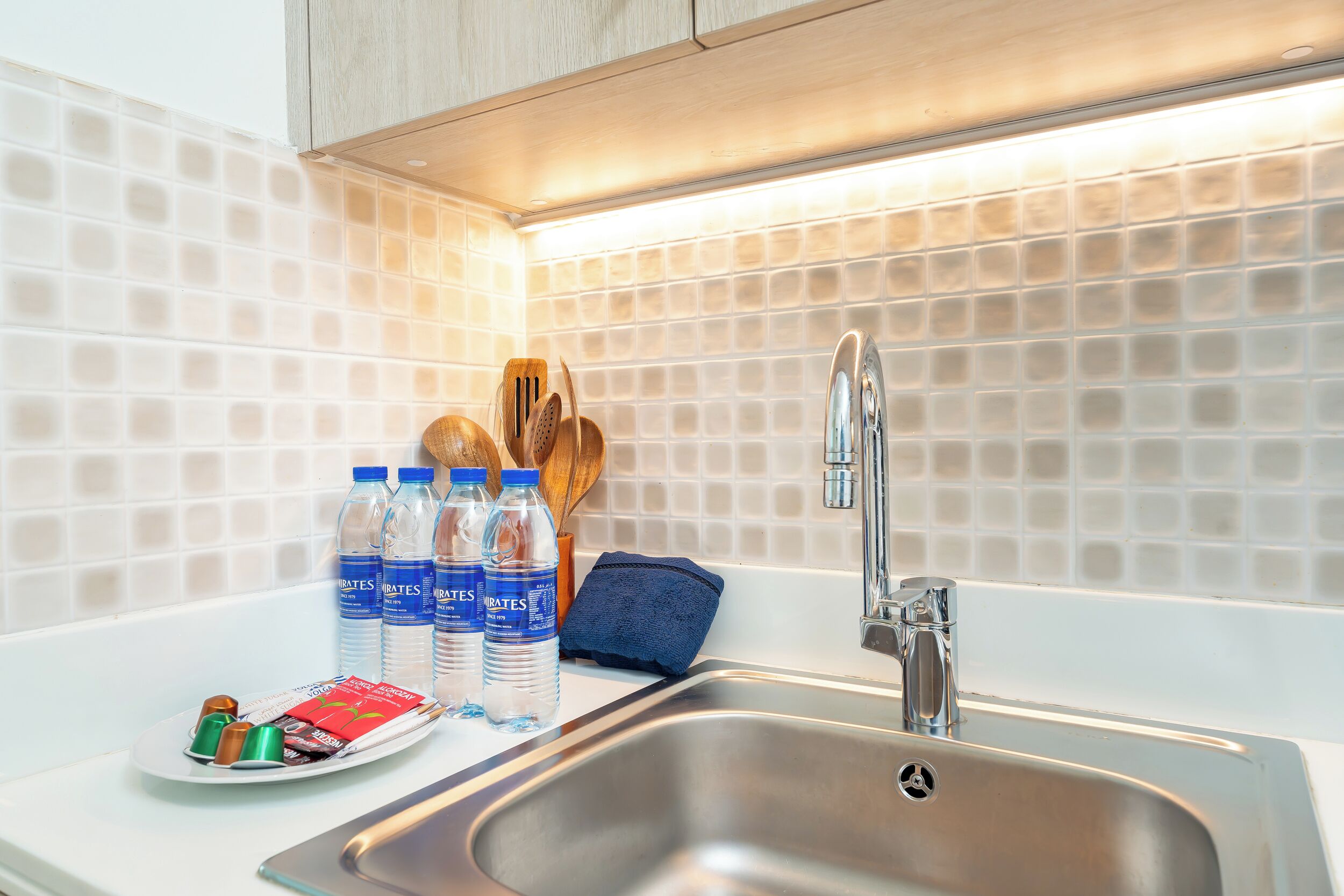Una cocina moderna con un lavamanos de acero inoxidable, salpicadero de azulejos blancos y utensilios de madera sobre la encimera.