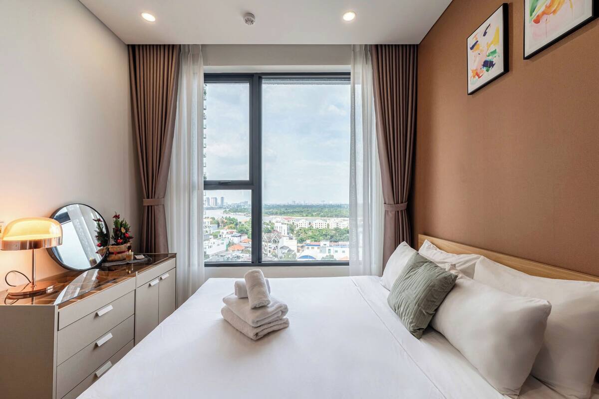 Quarto de hotel com uma janela grande, cama com lençóis brancos, mesa de cabeceira com abajur e um espelho.