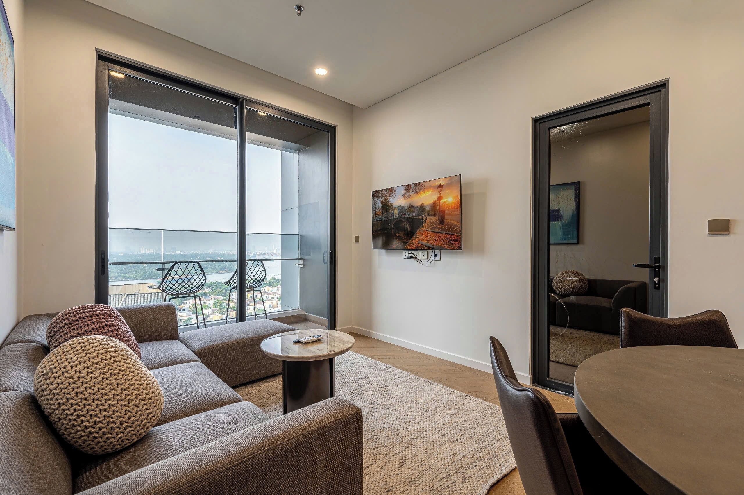 A modern living room with a sofa, round coffee table, and a dining table. Large windows offer a view of the city.