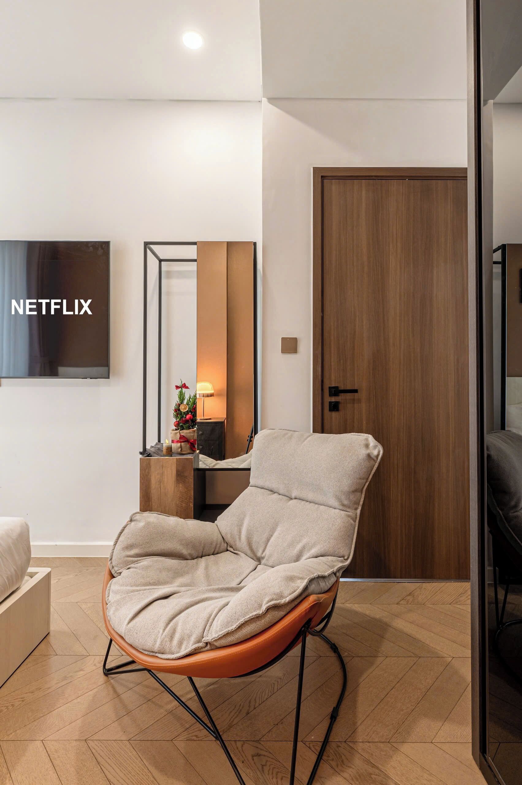 A modern living room with a beige armchair, a wooden door, a television mounted on the wall, and a small table with a lamp and decorative items.