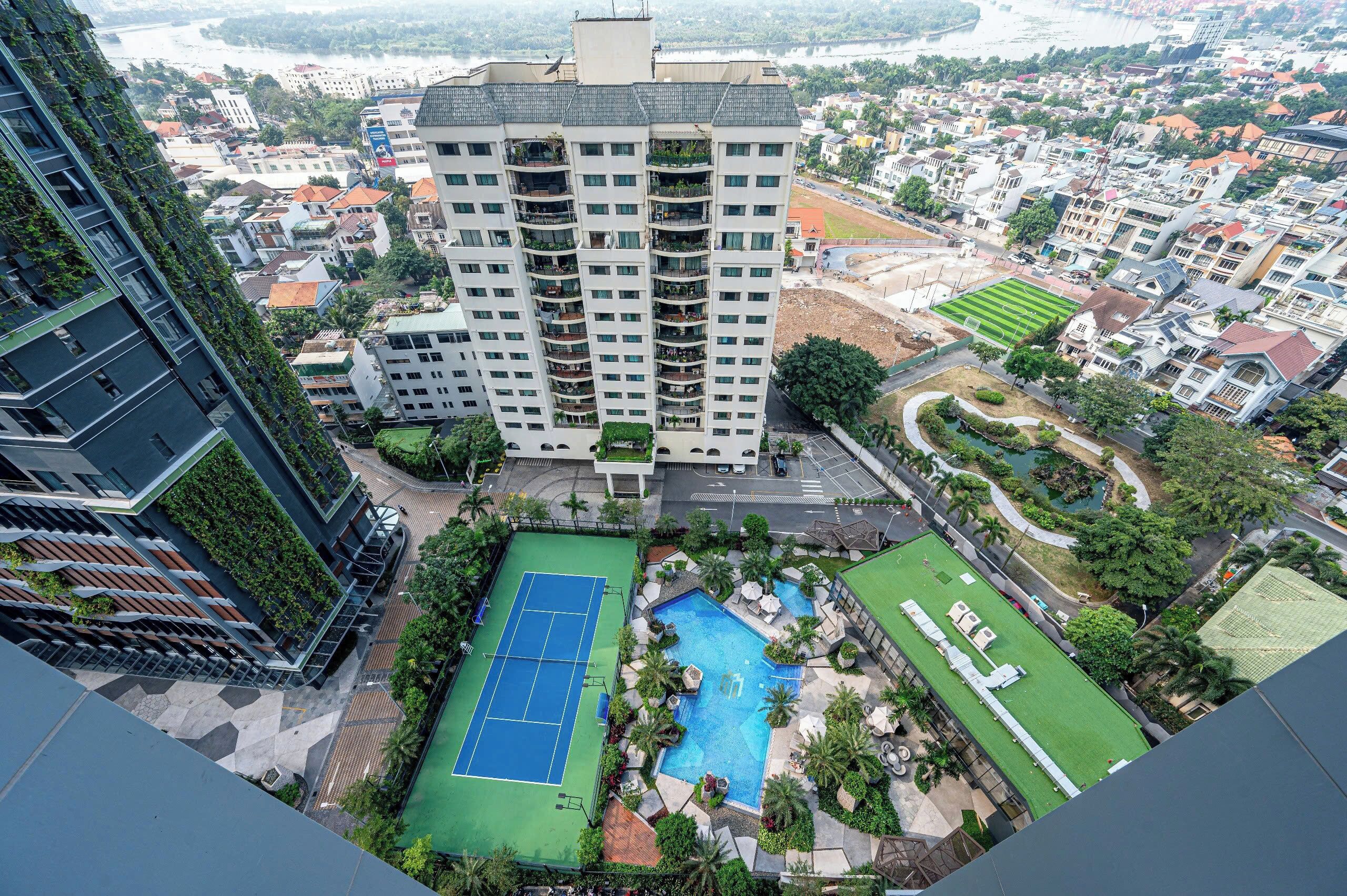 A high-rise building with a rooftop pool and tennis court, surrounded by greenery and residential buildings.