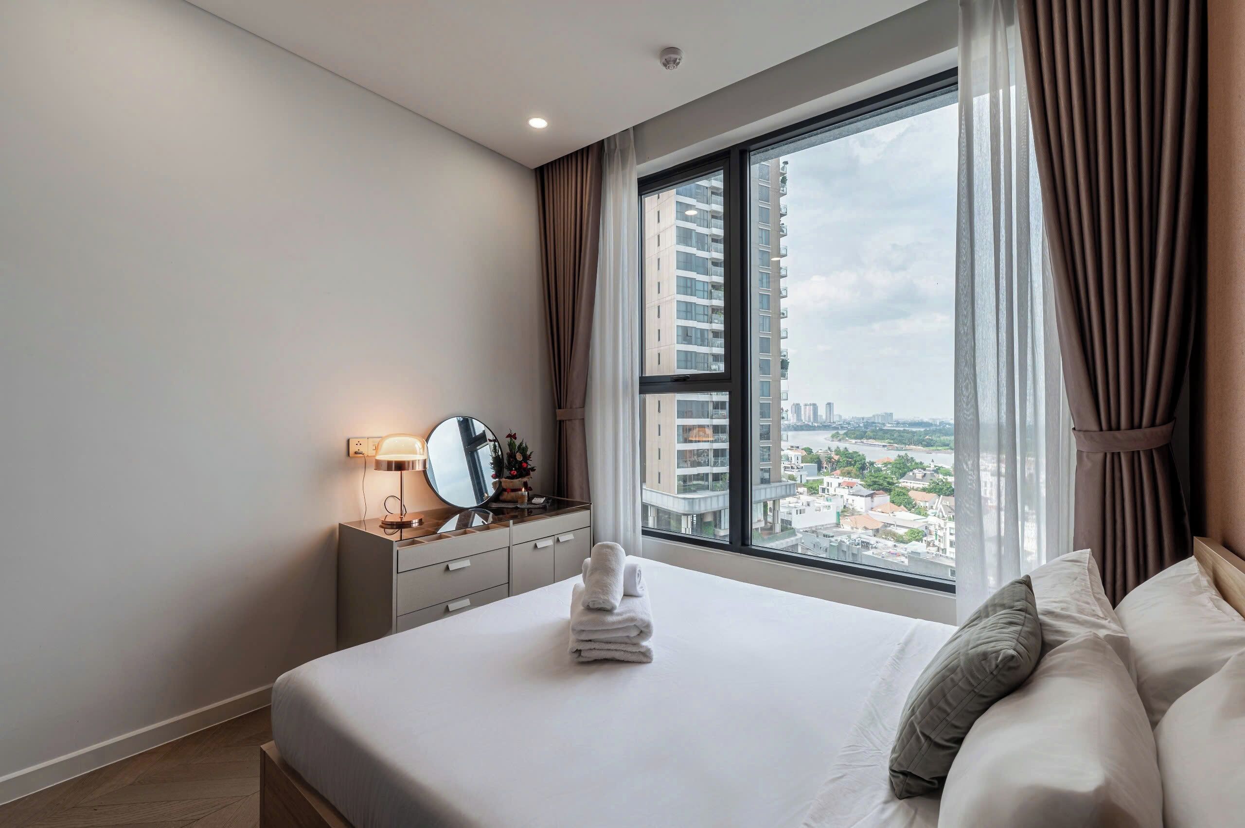 A hotel room with a large window, a bed with neatly folded towels, a bedside table with a lamp, and a mirror.