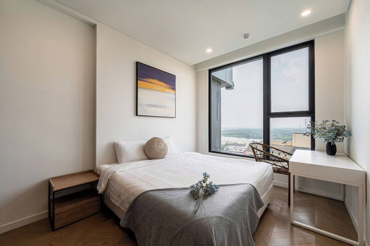 Quarto moderno com uma janela grande, uma cama com cobertor cinza, uma mesa de cabeceira de madeira e uma pequena escrivaninha com um vaso de flores.