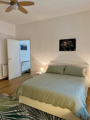 Ein Schlafzimmer mit Bett, Nachttisch, Deckenventilator, Heizkörper und einer Tür zu einem anderen Raum.