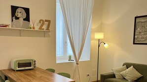 Een kamer met een houten eettafel, groene stoelen, een groene bank, een vloerlamp en een raam met witte gordijnen.