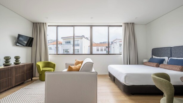 A modern hotel room with a large window, a bed, a sofa, a TV, and a small desk. - Aspasios Picaria Apartments (Porto)