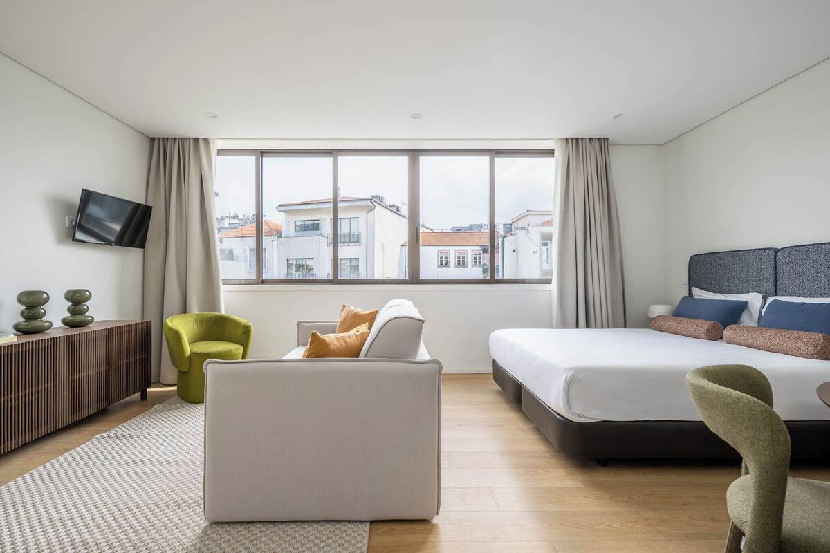 A modern hotel room with a large window, a bed, a sofa, a TV, and a small desk.