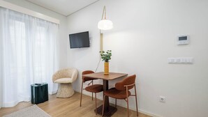 A modern living room with a wooden dining table, four chairs, a small side table, a floor lamp, a television, and a thermostat. - Aspasios Picaria Apartments (Porto)