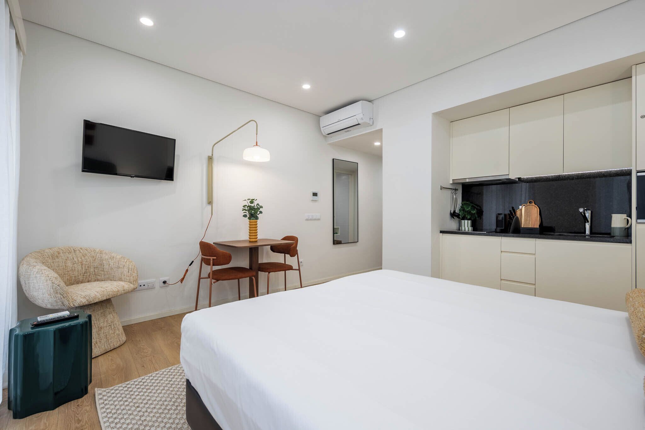 A modern hotel room with a bed, a desk with chairs, a flat-screen TV, an air conditioner, and a kitchenette.