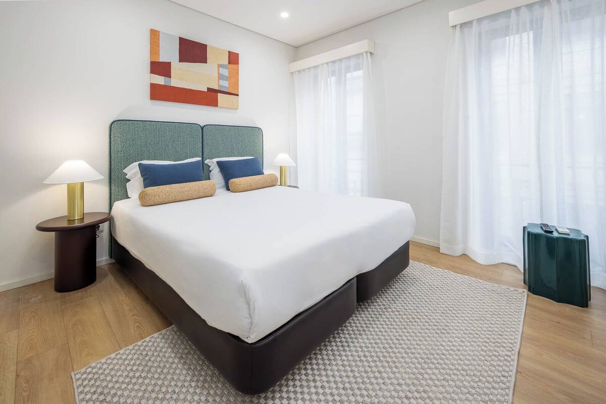 A modern hotel room with a large bed, two bedside tables with lamps, a small table with a remote control, and a geometric artwork on the wall.