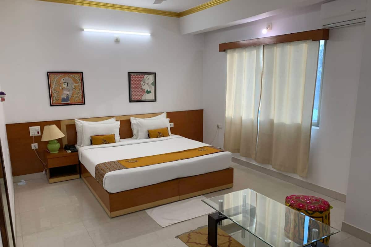 Una habitación de hotel con una cama grande, una mesita de noche con lámpara, una mesa de vidrio y una ventana con cortinas.