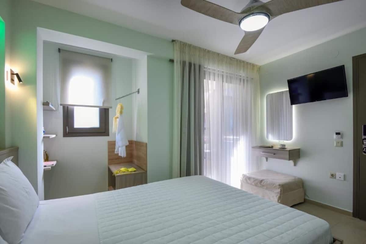 A hotel room with a bed, a ceiling fan, a TV, a window with curtains, and a small bathroom area.