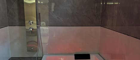 A modern bathroom with a glass-enclosed shower, a heated bathtub with red lighting, and marble walls.