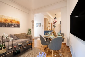 A modern living room with a sofa, coffee table, and dining area. There is a painting on the wall, a vase with flowers, and a TV mounted on the wall. - Miss H Haven (Athina)