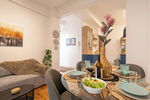A modern living room with a dining area, featuring a sofa, a glass table, and a vase with flowers. - Miss H Haven (Athina)