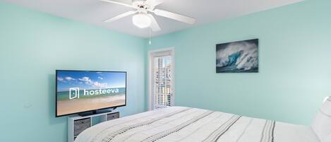 Ein Schlafzimmer mit Bett, Deckenventilator, Fernseher mit der Anzeige 'hosteeva', Fenster und einem Gemälde einer Welle.