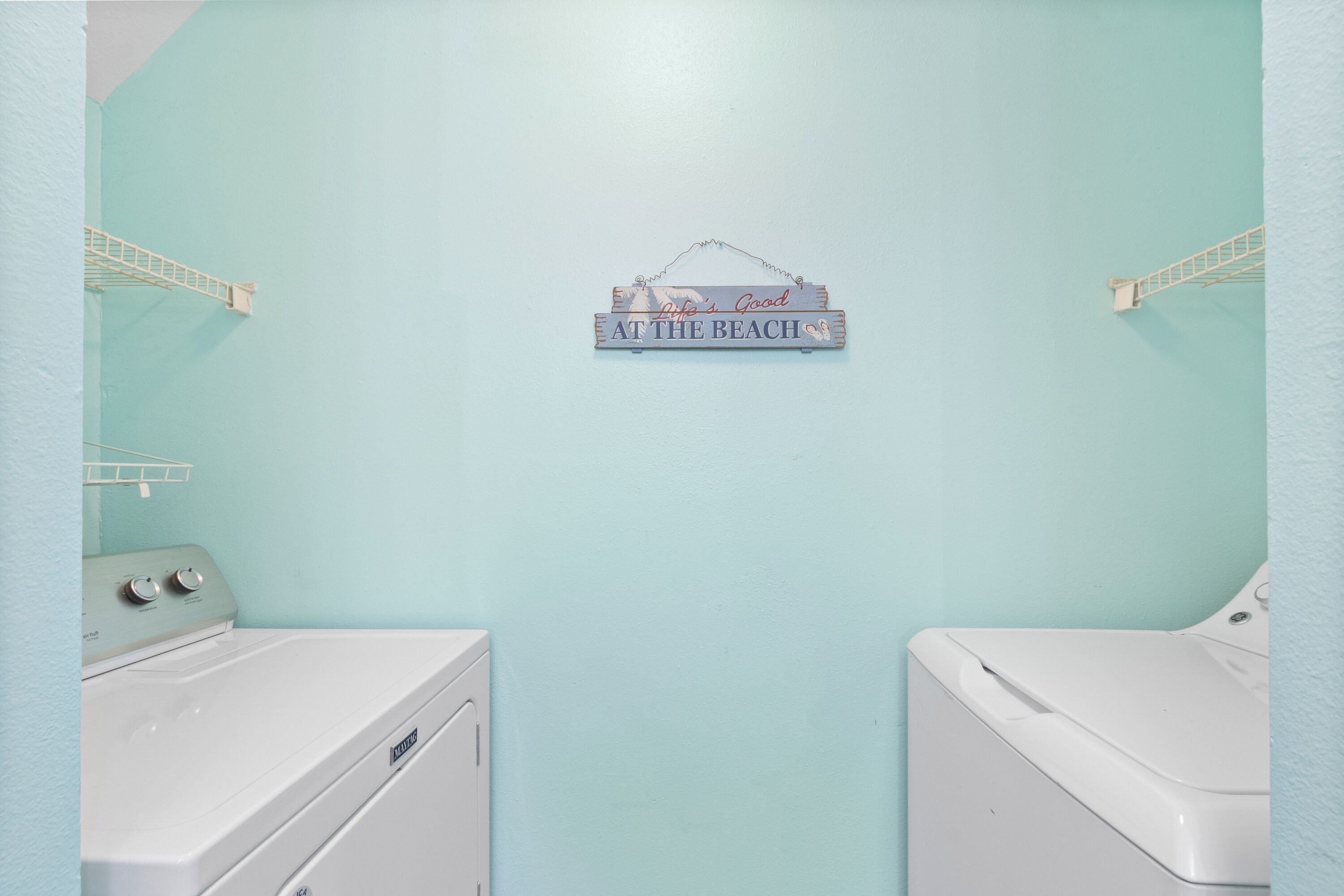 Un lavadero con dos lavadoras y secadoras blancas, una pared azul claro y un cartel que dice 'Life's Good at the Beach'.