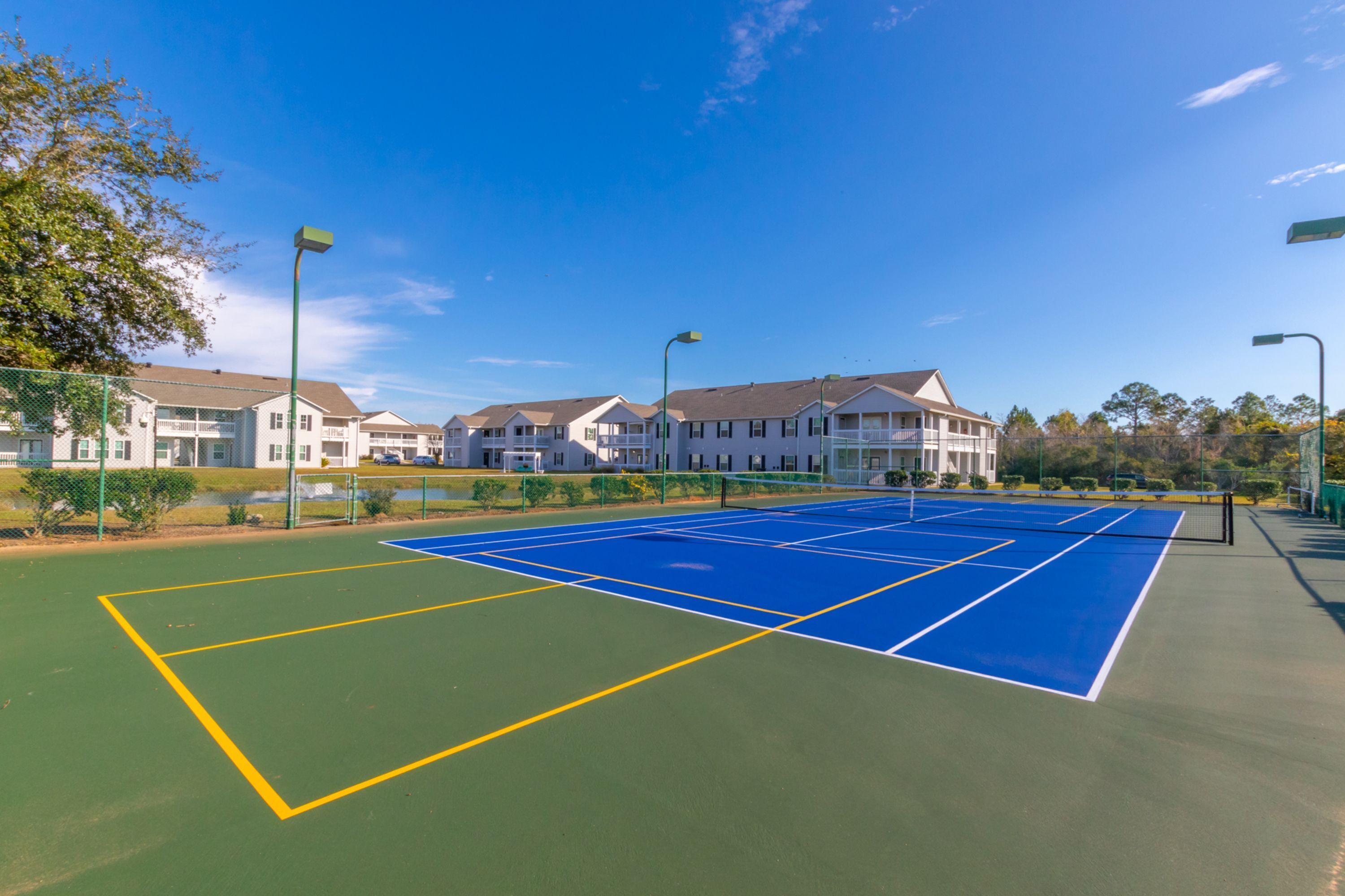 A tennis court with a net, surrounded by a fence, and adjacent to a row of white buildings with balconies.