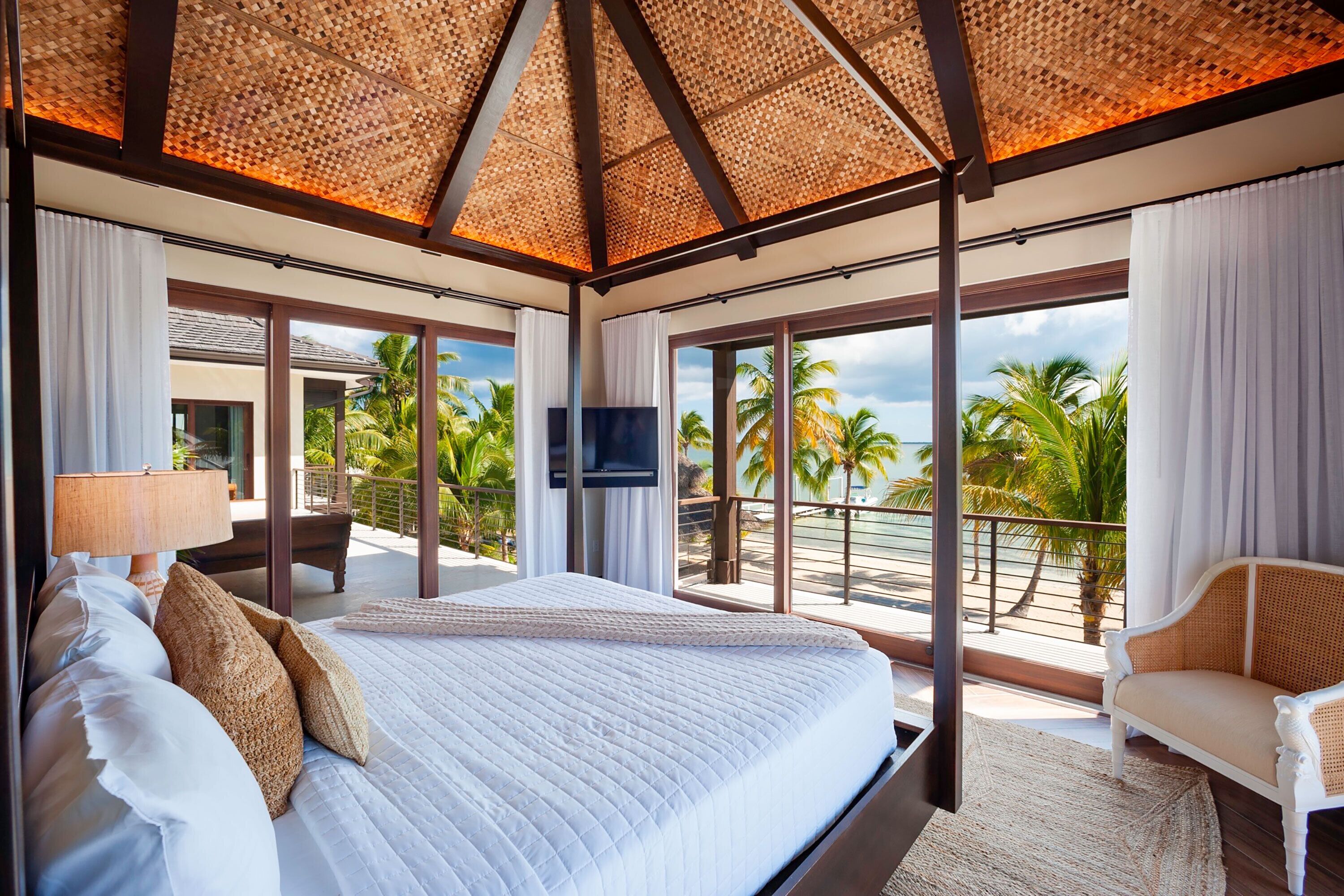 A spacious bedroom with a large bed, a television, and a balcony with a view of palm trees and the ocean.