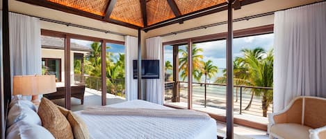 Un dormitorio espacioso con una cama grande, televisión y un balcón con vistas a palmeras y al océano.