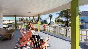 A covered outdoor area with wooden chairs and a wicker sofa, a white railing, and a view of palm trees and buildings in the background. - Casa Bella 4 Bedroom Home (Port Bolivar)