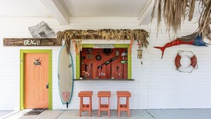 A beach-themed bar with a surfboard, three stools, and a sign reading "SAND BAR OPEN". - Casa Bella 4 Bedroom Home (Port Bolivar)